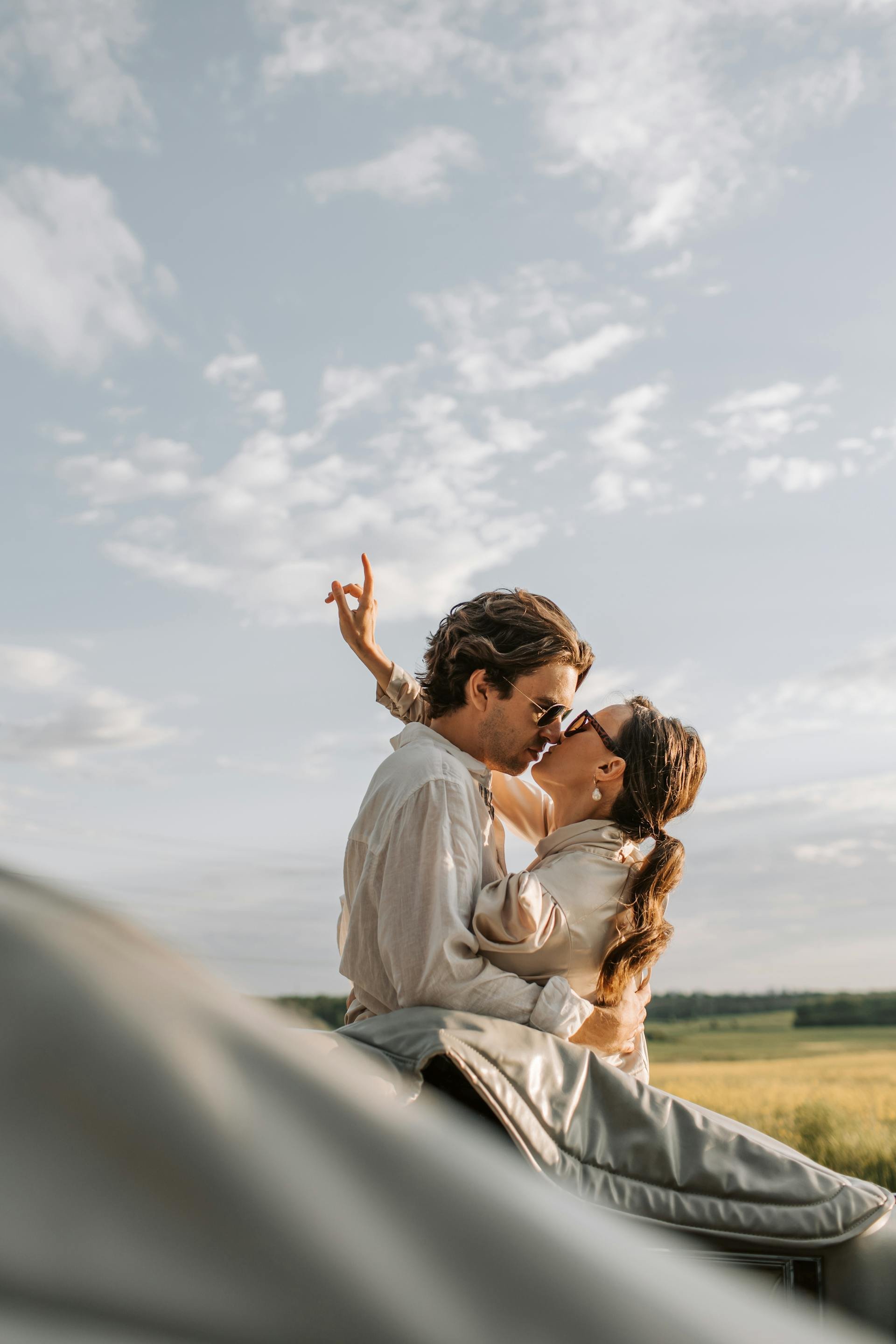 Wedding Website Design, Digital Wedding Stationery, Web Design and Branding for Wedding A couple embraces and kisses on a sunny day, with one person raising an arm in the air. They are sitting on a blanket in a grassy field under a partly cloudy sky.